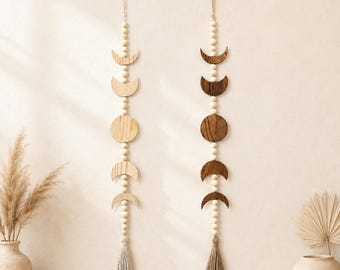 May include: Two vertical wall hangings featuring wooden moon phases and round beads. The left hanging is light wood with a light gray tassel, and the right hanging is dark wood with a dark brown tassel. Both have a loop at the top for hanging.