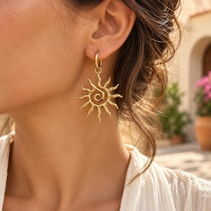 Peut inclure: Une boucle d'oreille dorée composée d'un anneau circulaire attaché à un pendentif texturé en forme de soleil. Le pendentif présente un motif en spirale centrale avec des rayons ondulés rayonnants, créant un aspect solaire détaillé.
