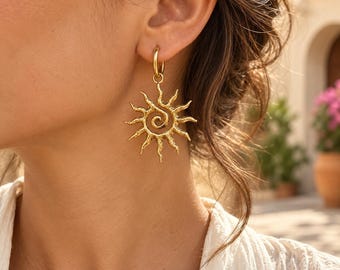 May include: A gold-toned earring featuring a circular hoop attached to a textured sun-shaped pendant. The pendant has a central spiral design with radiating wavy rays extending outward, creating a detailed celestial sunburst appearance.