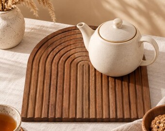 May include: A dark brown wooden serving tray with a curved arch shape and vertical grooved lines. A round, speckled off-white ceramic teapot with a matching lid sits on the tray. The tray features a flat base and a rounded top edge.