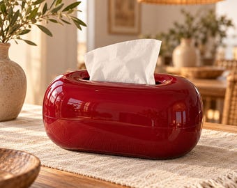 May include: A glossy, deep red, oval-shaped tissue box cover with a smooth, reflective surface. A single white paper tissue is pulled through the central elongated opening at the top. The container has a rounded, seamless design with a horizontal seam.