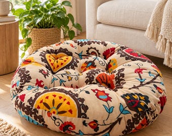 May include: A round floor cushion featuring a cream-colored base with a repeating pattern of dark brown, bright yellow, red, and light blue floral and paisley motifs. The cushion has a tufted surface with visible indentations and soft edges.