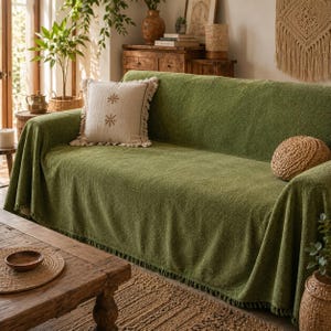 Peut inclure: Une housse de canapé vert olive texturée avec un ourlet à franges, accompagnée d'un coussin carré blanc cassé avec deux motifs brodés marron en forme de flocon et des bords frangés, ainsi qu'un coussin décoratif rond en osier beige.