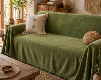 May include: A textured olive green sofa cover with a fringed hem, featuring a square off-white throw pillow with two embroidered brown snowflake-like motifs and fringed edges, alongside a round, woven, tan-colored decorative throw pillow.
