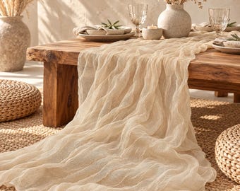 Puede incluir: Un camino de mesa largo y transparente de tela de queso beige con una textura arrugada, colocado sobre una mesa de madera. La tela es ligera y semitransparente, con pliegues suaves y fluidos que se extienden por la superficie de la mesa.