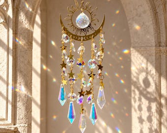 Puede incluir: Un atrapasoles dorado con forma de sol y luna creciente, que presenta una esfera de cristal transparente central, cadenas colgantes con pequeñas estrellas doradas y múltiples prismas de cristal iridiscente que refractan la luz.
