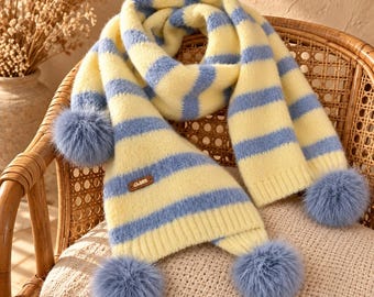 Puede incluir: Una bufanda de punto suave con rayas horizontales en amarillo pálido y azul bígaro. La bufanda está rematada con tres pompones redondos y esponjosos de color azul bígaro en los extremos e incluye una pequeña etiqueta marrón con texto.