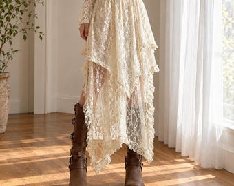 May include: A cream-colored, high-waisted, midi-length skirt crafted from floral lace fabric. The skirt features a layered, asymmetrical hemline with ruffled edges and a sheer, lightweight appearance, paired with brown leather mid-calf boots.