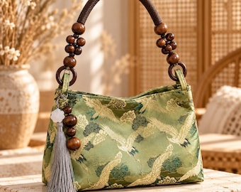 May include: A light green handbag featuring a repeating pattern of white cranes and dark green foliage. The bag has a dark brown woven handle accented with wooden beads and a decorative gray tassel charm with a white flower and wooden beads.