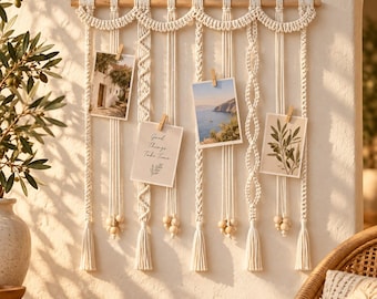 May include: A cream-colored macrame wall hanging suspended from a wooden dowel, featuring intricate knotted patterns, hanging cords with wooden beads, and tassels. Four rectangular photographs are attached to the cords with small wooden clips.