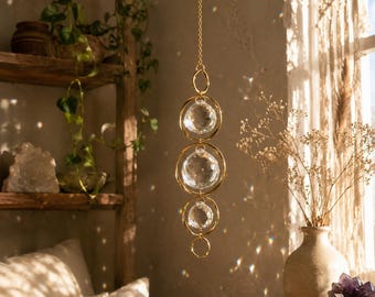 May include: A hanging suncatcher featuring three clear, faceted spherical crystals suspended within gold-toned metal rings. The ornament is attached to a delicate gold-toned chain, designed to catch and refract light, creating small rainbow reflections.