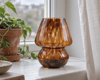 May include: A mushroom-shaped table lamp crafted from translucent amber-colored glass. The lamp features a wide, flared shade and a rounded base, both displaying a distinct, swirling, light-catching pattern throughout the glass material.