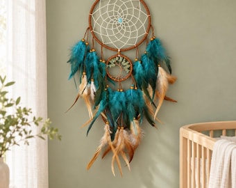 May include: A dream catcher featuring a large brown hoop with a white web and a central turquoise bead. Below, a smaller hoop contains a wire tree sculpture with green stone leaves. Teal and tan feathers hang from both hoops with gold beads.