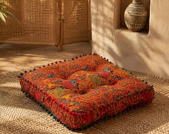May include: A square floor cushion featuring a vibrant orange and red fabric with a detailed multi-colored floral and geometric pattern. The cushion is tufted in the center and finished with small, dark-colored pom-pom trim along all four edges.