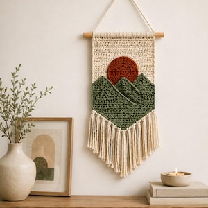 Op de afbeelding: Een wandkleed met een gehaakt landschapsontwerp met een crèmekleurige achtergrond, een donkeroranje cirkelvormige zon en donkergroene bergtoppen. Het stuk hangt aan een houten stok met een crèmekleurig koord en crèmekleurige franjes.