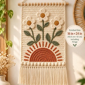 Könnte beinhalten: Ein cremefarbener Häkel-Wandbehang mit einer rostfarbenen Halbkreis-Sonne, drei weißen Blumen mit gelben Mitten und grünen Stielen. Er enthält einen Holzstab, eine Reihe von Noppenmaschen und Fransen. Die fertige Größe beträgt 40,6 cm x 61 cm.