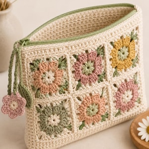 May include: A cream-colored crochet pouch featuring six square panels with floral motifs in shades of dusty pink, sage green, and mustard yellow. The pouch has a sage green zipper closure with a small matching flower and leaf charm attached.