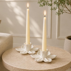 May include: Two white ceramic candle holders shaped like open flowers, each featuring a central cylindrical socket holding a tall, slender, cream-colored taper candle with a lit flame, resting on a circular surface.