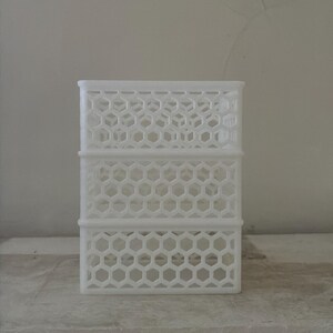 May include: A white rectangular storage container featuring a repeating hexagonal honeycomb pattern across its sides. The structure is divided into three horizontal sections by solid white bands, creating a grid-like appearance for the open-work design.