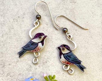 May include: A pair of silver earrings featuring hand-painted enamel bird charms. The birds are black, white, and brown with a purple head and a detailed design. The earrings have a silver hook closure.
