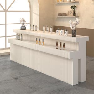 May include: A white, tiered reception desk featuring a vertical fluted texture on its front and side panels. The desk has two levels for display, holding various small glass bottles with white labels and two clear vases containing white flowers.