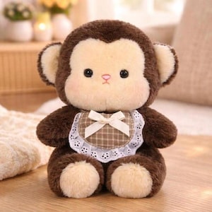May include: A plush monkey toy with dark brown fur and a cream-colored face. The monkey features black eyes, a small pink nose, and rounded ears. It wears a brown and white checkered bib with a white lace trim and a small cream-colored ribbon bow.