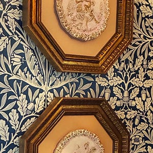 May include: Two octagonal wall frames with gold-toned borders and light-colored backings. Each frame contains a central oval relief plaque featuring a raised, textured scene of figures in a garden setting with intricate, ornate detailing.
