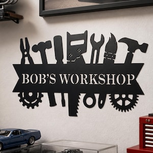 Puede incluir: Un letrero de pared de metal negro con el texto BOB'S WORKSHOP centrado en una pancarta. El diseño incluye siluetas de varias herramientas como alicates, un martillo, un destornillador, una sierra, una llave inglesa y dos engranajes.