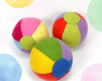 May include: Three colorful felt balls in shades of pink, yellow, red, green, purple, and blue. Each ball is segmented with contrasting colors and a circular patch. The balls are arranged on a white background, suggesting a playful theme.