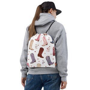 Può includere: Uno zaino bianco con chiusura a coulisse caratterizzato da un motivo ripetuto di stivali da cowboy di vari colori, tra cui rosa, viola, marrone e rosso, intervallati da piccole illustrazioni floreali e stelle, con spallacci a corda neri.