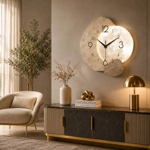 May include: A wall clock with an irregular, stone-like beige frame and a circular, illuminated moon-textured face. The clock features black Arabic numerals and thin black hands. It is mounted on a wall above a sideboard with a dark marble top.