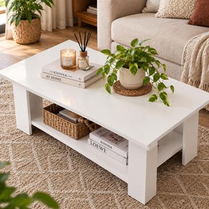 May include: A rectangular white coffee table with a smooth top surface and a lower storage shelf. The table features thick legs and holds a potted green plant, a candle, a reed diffuser, and stacked books titled The Kinfolk Home and Loewe.