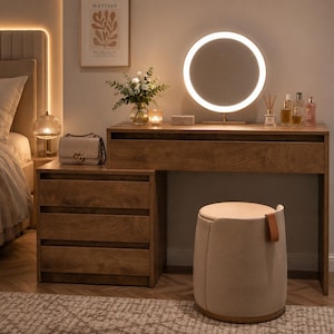 May include: A wooden vanity table with three drawers on the left side and a long drawer across the top. A circular LED mirror sits on the surface next to a vase of flowers and perfume bottles. A beige upholstered stool with a leather pull tab sits below.