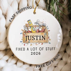 May include: A round white ceramic ornament featuring a watercolor illustration of a wooden toolbox filled with tools, including a hammer, level, and pliers. The text reads JUSTIN, FIXED A LOT OF STUFF, and 2026, surrounded by colorful string lights.