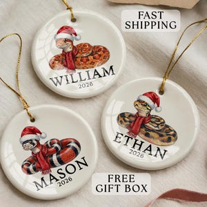 May include: Three round white ceramic ornaments featuring illustrations of snakes wearing red Santa hats and scarves. Each ornament is personalized with a name, William, Ethan, or Mason, and the year 2026 printed in black text.