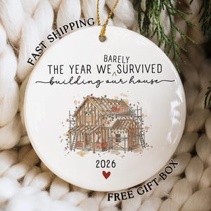 May include: A round white ceramic ornament featuring a watercolor illustration of a house under construction with scaffolding and string lights. The text reads BARELY THE YEAR WE SURVIVED building our house and 2026 with a small red heart below.