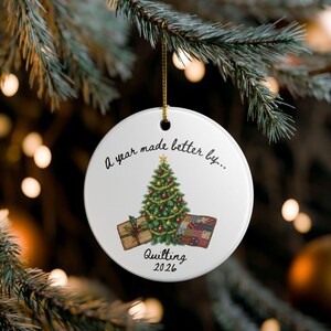 May include: A round white ceramic ornament hanging by a gold string. The front features a printed illustration of a decorated Christmas tree flanked by a wrapped gift and a colorful quilt, with the text A year made better by... Quilting 2026.