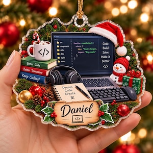 Op de afbeelding: Een decoratief acrylen ornament met een laptop die code weergeeft, een kerstmuts, een sneeuwpop, een mok met een codesymbool, gestapelde boeken, een notitieblok met Dream Code Create en een naamplaatje met de tekst Daniel.