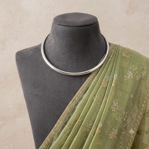 May include: A smooth, polished silver-toned metal collar necklace with a circular, rigid design. The necklace is displayed on a dark grey mannequin bust draped with a sheer, light olive green fabric featuring delicate gold-toned floral embroidery.