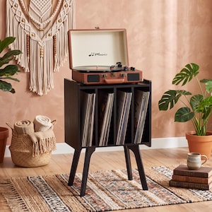 May include: A black wooden record storage cabinet with four vertical compartments for vinyl records, standing on four tapered legs. A brown portable suitcase-style record player with the text Music is placed on top of the storage unit.