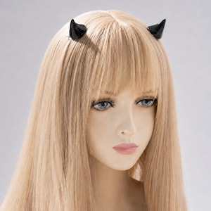 Puede incluir: Un par de pequeños cuernos de diablo negros y brillantes unidos a un accesorio para el cabello. Los cuernos son lisos, curvos y puntiagudos, colocados simétricamente sobre una cabeza de cabello rubio liso con flequillo y ojos azules.