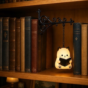 May include: A black metal wall-mounted bracket with ornate scrollwork supports a small, glowing, cream-colored cat-shaped lamp. The cat figure is holding a small black book and is suspended from the bracket by a short black metal chain.