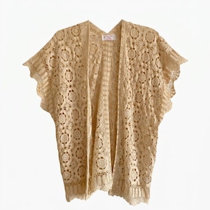 May include: A beige open-front cardigan featuring an intricate crochet pattern with circular floral motifs and scalloped edges. The garment has short, wide sleeves and a straight hemline, with a small white rectangular label at the inner neckline.