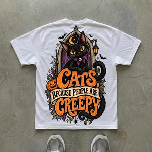 May include: A white t-shirt featuring a graphic on the back of a black cat with yellow eyes and a purple bow tie, positioned in front of a gothic archway. The text reads "CATS BECAUSE PEOPLE ARE CREEPY" in orange and white with pumpkin and bat accents.