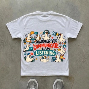 Puede incluir: Una camiseta blanca con un gráfico colorido de patos de dibujos animados realizando diversas actividades de comunicación. El texto central dice: "HOWEVER YOU COMMUNICATE, I AM LISTENING." El diseño incluye patos con ordenadores portátiles, auriculares y megáfonos.