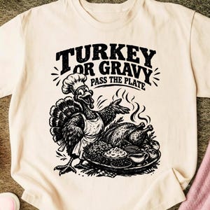 Może przedstawiać: Kremowy t-shirt z czarnym nadrukiem graficznym przedstawiającym indyka w czapce kucharskiej i fartuchu. Indyk wskazuje na półmisek z jedzeniem. Napis głosi "TURKEY OR GRAVY" oraz "PASS THE PLATE" pogrubioną, stylizowaną czcionką.