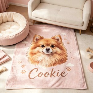 May include: A soft, rectangular, light pink fleece blanket featuring a large, detailed illustration of a fluffy, tan Pomeranian dog face in the center. The blanket includes small paw print graphics and the name Cookie written in cursive script.