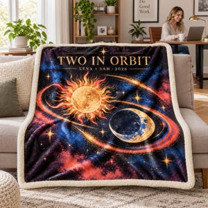 May include: A plush throw blanket with a cream-colored sherpa lining and a dark space-themed print featuring a glowing sun and crescent moon. The text reads TWO IN ORBIT, LENA + SAM, 2026, surrounded by swirling orange rings and stars.