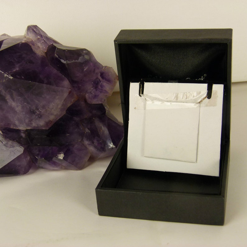 May include: A black jewelry box with an open lid, revealing a white card. A large, rough-cut amethyst crystal is to the left. The box is empty, suggesting it is for display or storage of jewelry.