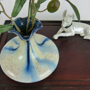 May include: A white ceramic vase with blue and brown glaze, featuring a ruffled rim and a narrow neck. The vase is filled with a sprig of green leaves and a single olive.